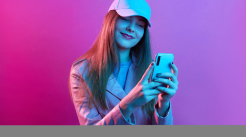 Portrait of young beautiful fashionable model wearing leather baker and baseball cap, holding smart phone in hands, texting with friends or checking social networks.