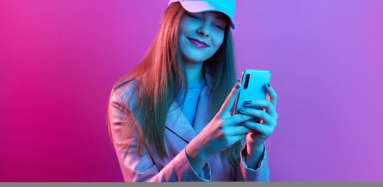 Portrait of young beautiful fashionable model wearing leather baker and baseball cap, holding smart phone in hands, texting with friends or checking social networks.
