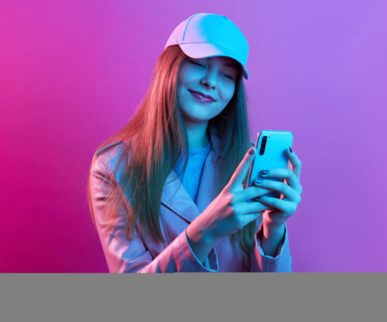 Portrait of young beautiful fashionable model wearing leather baker and baseball cap, holding smart phone in hands, texting with friends or checking social networks.